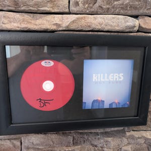 Puede incluir: CD enmarcado y portada del álbum "Hot Fuss" de The Killers. El CD es rojo con una firma y una pegatina de autenticación PSA. La portada presenta el nombre de la banda y un paisaje urbano.