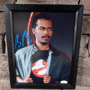 May include: Framed photograph of an actor wearing a black t-shirt with the Ghostbusters logo. The photo is in a black frame and is signed in blue ink. The actor is wearing a light blue jacket over the t-shirt.