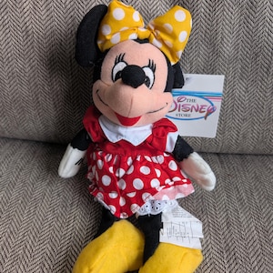 Puede incluir: Un peluche de Minnie Mouse. La mu&ntilde;eca tiene un vestido rojo con lunares blancos, un lazo amarillo con lunares blancos y zapatos amarillos. La etiqueta dice "The Disney Store".