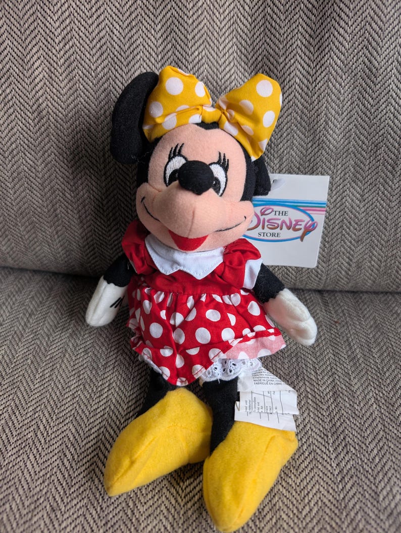 Puede incluir: Un peluche de Minnie Mouse. La mu&ntilde;eca tiene un vestido rojo con lunares blancos, un lazo amarillo con lunares blancos y zapatos amarillos. La etiqueta dice "The Disney Store".