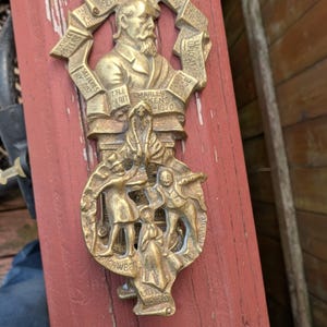 May include: Brass door knocker featuring a relief sculpture of Charles Dickens and characters from his novels. The knocker is shaped like a book with the title "Little Dorrit" and the text "Charles Dickens 1870" on the front. The back of the knocker features a scene from the novel "Our Mutual Friend" with the text "Our Mutual Friend" and "The Gaieties" on the bottom.