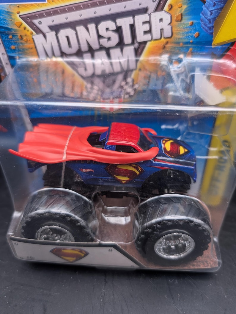 May include: A blue and red Monster Jam truck toy featuring the Superman logo. The truck has large black tires and a red cape. The packaging includes the words "Monster Jam" and "Off Road".