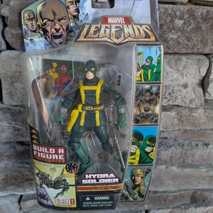 May include: A green and yellow Marvel Legends Hydra Soldier action figure with a gun and a snake. The figure is part of a Build a Figure collection. The packaging includes the text "Build a Figure Collection" and "Hydra Soldier".