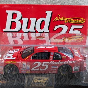 Op de afbeelding: Een rode Budweiser raceauto met nummer 25 aan de zijkant, in een transparante vitrine met een rode doos erbovenop waarop "Bud" en "Wally Dallenbach 25" staat.