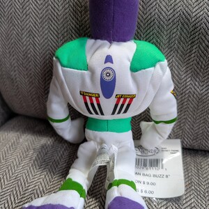 Op de afbeelding: Een pluche Buzz Lightyear pop uit de Disney-film Toy Story. De pop is wit met groene en paarse accenten en heeft de woorden "DANGER!" en "JET EXHAUST" op de rug. De pop is ongeveer 20 cm hoog.
