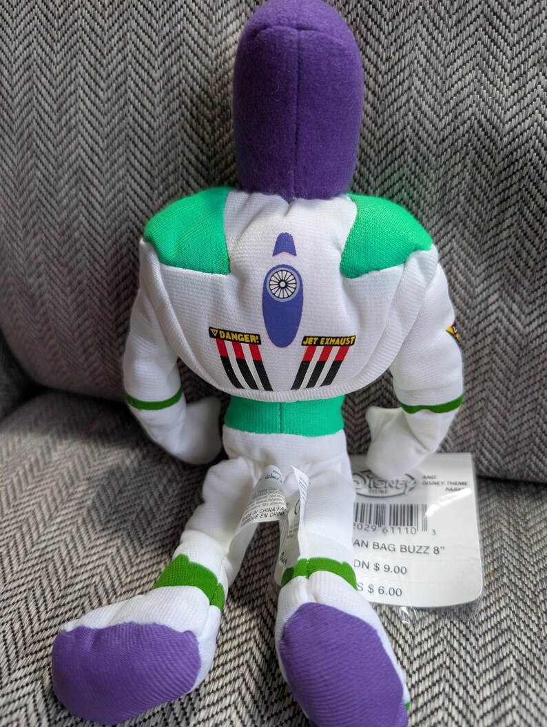 Op de afbeelding: Een pluche Buzz Lightyear pop uit de Disney-film Toy Story. De pop is wit met groene en paarse accenten en heeft de woorden "DANGER!" en "JET EXHAUST" op de rug. De pop is ongeveer 20 cm hoog.