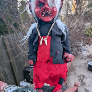 May include: A red and black demon doll with white hair and horns. The doll is wearing a black robe and a red skirt. The doll is standing on a wooden platform.