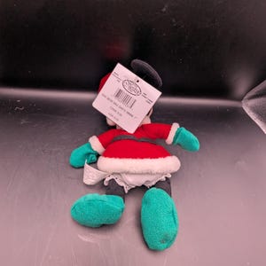 K&ouml;nnte beinhalten: Eine Pl&uuml;schpuppe von Minnie Mouse in einem Weihnachtsmann-Outfit. Die Puppe hat ein rotes Kleid mit wei&szlig;em Besatz, gr&uuml;ne Handschuhe und Schuhe sowie einen schwarzen Kopf mit einer Schleife. Ein Etikett ist an der Puppe befestigt.