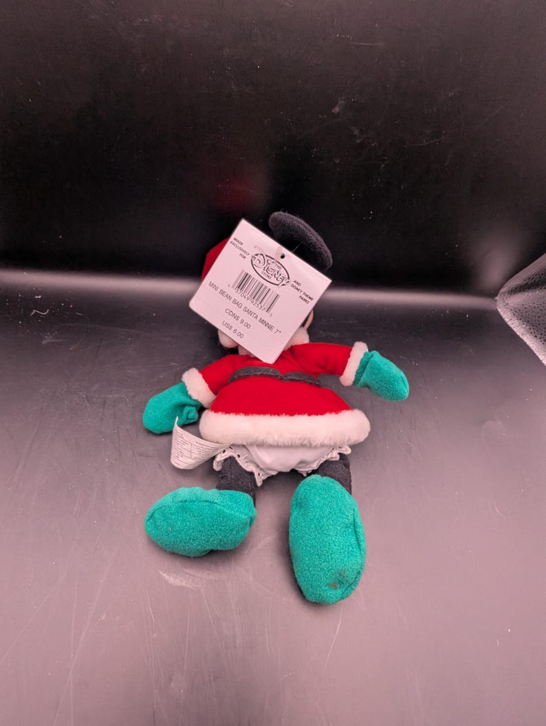 K&ouml;nnte beinhalten: Eine Pl&uuml;schpuppe von Minnie Mouse in einem Weihnachtsmann-Outfit. Die Puppe hat ein rotes Kleid mit wei&szlig;em Besatz, gr&uuml;ne Handschuhe und Schuhe sowie einen schwarzen Kopf mit einer Schleife. Ein Etikett ist an der Puppe befestigt.