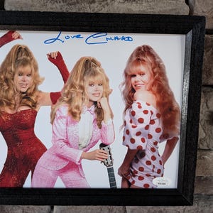 May include: A framed photograph of three women with blonde hair wearing different outfits. The women are smiling and looking at the camera. The photo is signed "Love Emma".
