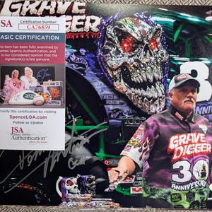 May include: A photograph featuring a man wearing a Grave Digger 30th Anniversary shirt and hat. The background includes a large skull graphic and the Grave Digger monster truck. A JSA authentication sticker is visible.