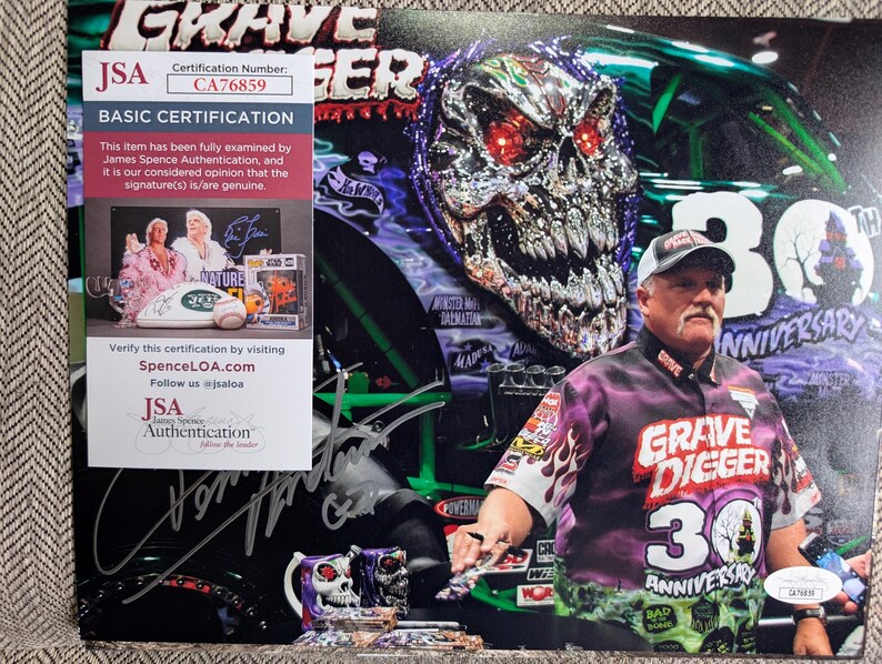 May include: A photograph featuring a man wearing a Grave Digger 30th Anniversary shirt and hat. The background includes a large skull graphic and the Grave Digger monster truck. A JSA authentication sticker is visible.