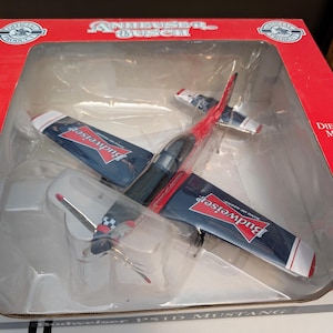 May include: A red, white, and blue model airplane with "Budweiser" printed on the side. The airplane is in a clear plastic box with a red and white background. The box says "Budweiser Busch" on the top.