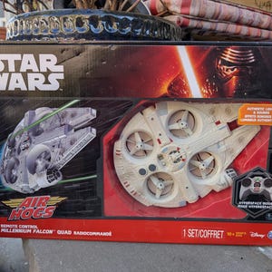 May include: A Star Wars Air Hogs remote control Millennium Falcon quadcopter toy. The toy is white with red and green accents. The box features a picture of the toy, the Star Wars logo, and the words "Air Hogs" and "Remote Control Millennium Falcon".