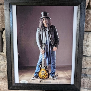 May include: A black and white photograph of a musician wearing a black leather jacket, a black top hat, sunglasses, and blue jeans. He is holding a brown and black electric guitar. The photograph is signed with the musician's name.