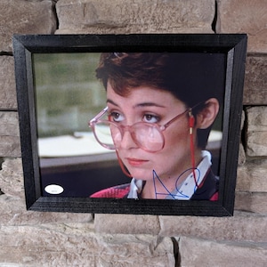 May include: Framed photograph of a person wearing pink-rimmed glasses, with a signature in blue ink. The photograph is set in a black wooden frame and is displayed against a textured stone wall.