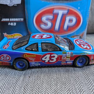 Op de afbeelding: Een blauwe, rode en paarse gegoten raceauto met nummer 43 en STP- en Cheerios-logo's. De auto is een Hot Wheels Crew's Choice model van John Andretti's auto.