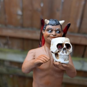 May include: A figurine of a devil-like figure holding a white skull. The figure has horns, red wings, and a stern expression. The figurine is painted in shades of brown and tan.
