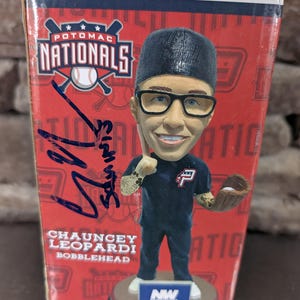 May include: A Potomac Nationals bobblehead of Chauncey Leopardi, wearing a black cap, glasses, and a navy blue uniform. The box is red with the team logo and a signature. The bobblehead is on a white base.