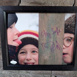 May include: Framed photograph featuring three children, each partially visible. One child sticks out their tongue, another smiles, and the third wears glasses. The words "Stay Safe" and a signature are visible on a wooden post. The frame is black.