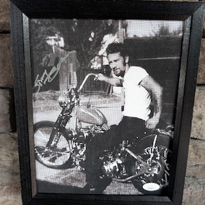 May include: Black and white framed photograph of a man on a motorcycle. The man is wearing a white t-shirt and dark trousers. The photograph is signed. The frame is black.