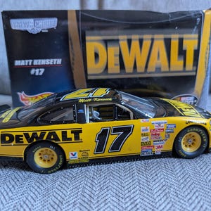 Op de afbeelding: Een geel en zwart model raceauto met nummer 17 en het "DEWALT" logo. De auto is een replica van Matt Kenseths auto, met sponsorlogo's. De verpakking toont het Hot Wheels logo.