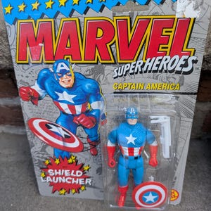 Puede incluir: Figura de acci&oacute;n del Capit&aacute;n Am&eacute;rica en su embalaje original. El embalaje dice "MARVEL SUPER HEROES" y presenta una ilustraci&oacute;n del Capit&aacute;n Am&eacute;rica. La figura es azul, roja y blanca, con un lanzador de escudo.