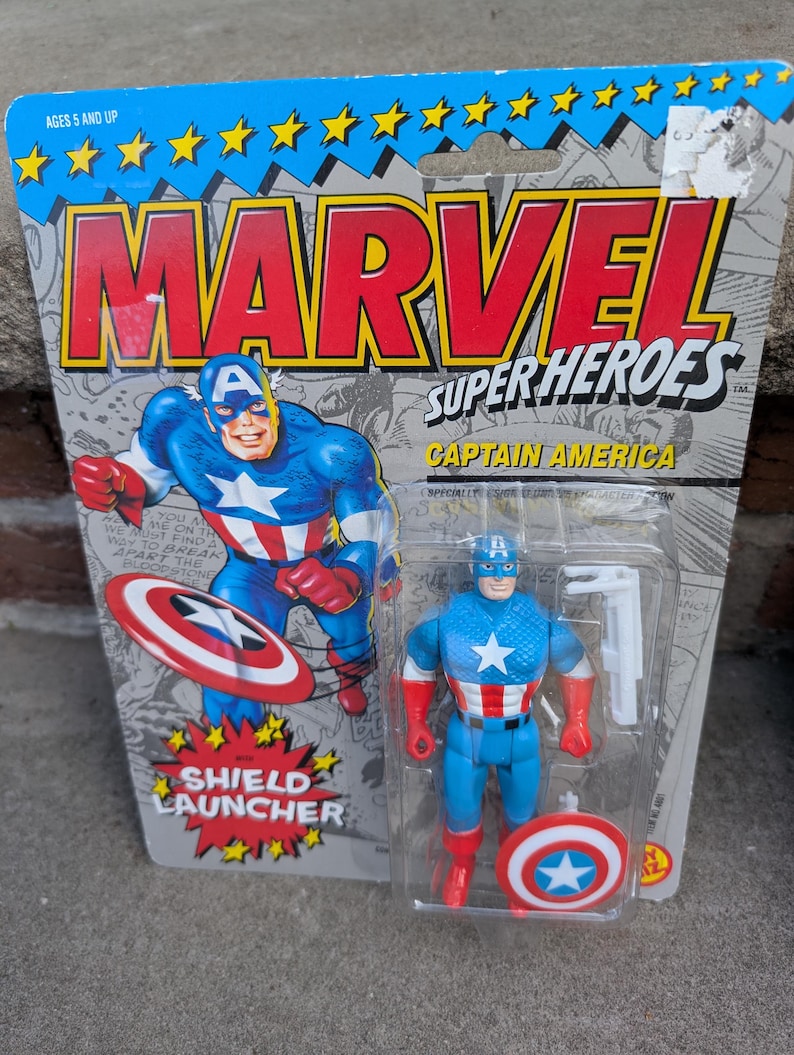 Puede incluir: Figura de acci&oacute;n del Capit&aacute;n Am&eacute;rica en su embalaje original. El embalaje dice "MARVEL SUPER HEROES" y presenta una ilustraci&oacute;n del Capit&aacute;n Am&eacute;rica. La figura es azul, roja y blanca, con un lanzador de escudo.