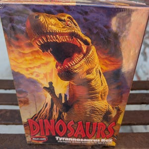 Könnte beinhalten: Eine Schachtel für ein Tyrannosaurus Rex Dinosaurier-Modellbausatz. Die Schachtel zeigt eine farbenfrohe Illustration eines brüllenden Tyrannosaurus Rex vor einem feurigen orange-gelben Hintergrund. Der Text auf der Schachtel lautet "Dinosaurs Tyrannosaurus Rex".