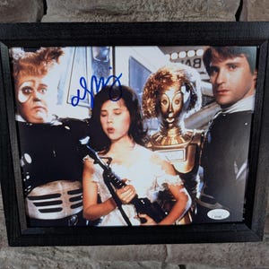 May include: Framed photograph featuring a group of people, including a robot, from a science fiction movie. The photo is signed in blue ink. The frame is black and rectangular, with a dark wood grain.