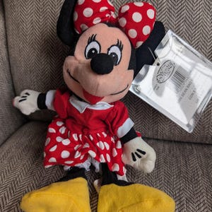 K&ouml;nnte beinhalten: Ein Minnie Mouse Mini-Beanbag-Pl&uuml;schtier. Das Pl&uuml;schtier hat ein rotes Kleid mit wei&szlig;en Punkten, eine passende Schleife und gelbe Schuhe. Das Spielzeug hat ein Etikett.