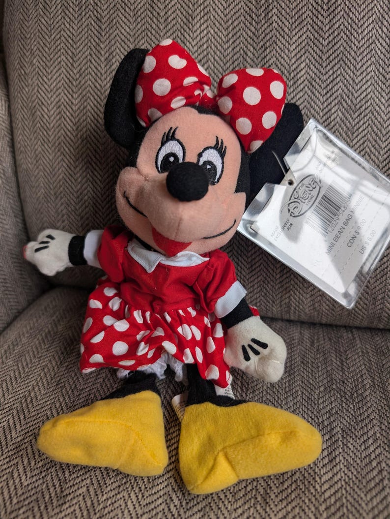 K&ouml;nnte beinhalten: Ein Minnie Mouse Mini-Beanbag-Pl&uuml;schtier. Das Pl&uuml;schtier hat ein rotes Kleid mit wei&szlig;en Punkten, eine passende Schleife und gelbe Schuhe. Das Spielzeug hat ein Etikett.