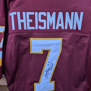 Pode incluir: Camisola de futebol grená com o nome "THEISMANN" em letras brancas no peito. O número "7" está na frente, contornado em dourado, com uma assinatura e a inscrição "P2 MVP".