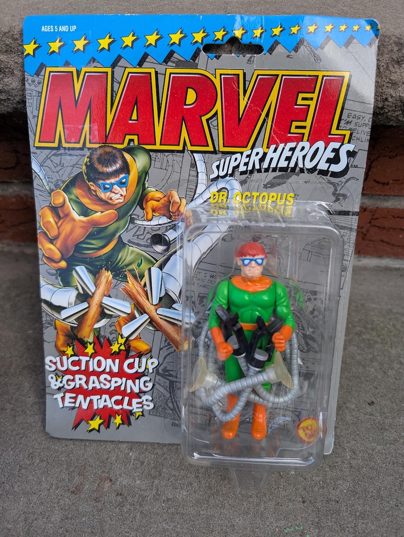 May include: A Marvel Super Heroes Dr. Octopus action figure in its original packaging. The figure is green and orange with four mechanical tentacles. The packaging features the Marvel logo and comic book-style artwork.