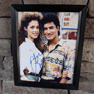 May include: Framed photograph featuring two smiling people, likely celebrities, with autographs. The frame is black, and the background is a stone wall. The photograph shows a man in a patterned shirt and a woman in a white top.