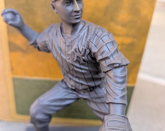 May include: A gray figurine of a baseball pitcher in a pinstriped uniform and cap, with the New York Yankees logo. The figure is in a throwing pose, holding a baseball and wearing a glove.