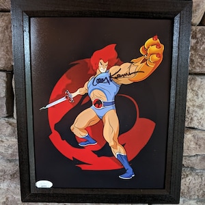 May include: A framed print of Lion-O, the main character from the ThunderCats cartoon, with a red and black background. The print is signed by the artist, Larry Kenney, who voiced Lion-O in the original cartoon.