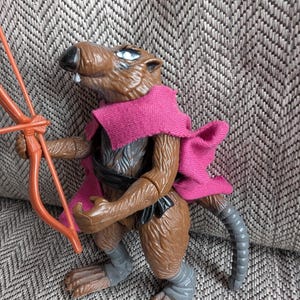 May include: A brown rat action figure with a pink cape, holding a brown and orange bow and arrow. The figure has a black belt and gray leg wraps.