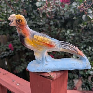 May include: A colorful bird statue with a blue base, perched on a red wooden post. The bird features yellow, orange, black, and blue paint. The statue is outdoors with a blurred green and pink floral background.