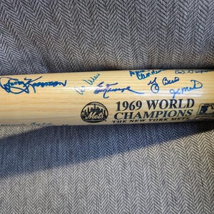 Pode incluir: Um taco de beisebol de madeira assinado pelos campeões da World Series dos New York Mets de 1969. O taco é marrom com letras azuis que dizem "1969 World Champions The New York Mets".
