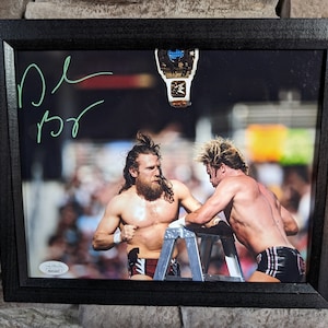 Könnte beinhalten: Ein Schwarzweißfoto von zwei Männern, die auf einer Leiter kämpfen. Das Foto ist mit "D.B.R." signiert und zeigt oben rechts einen Wrestling-Gürtel.