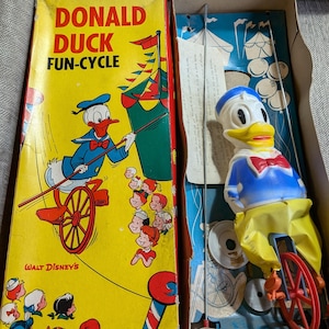 Może przedstawiać: Zabawka Donald Duck Fun-Cycle w stylu vintage w oryginalnym pudełku. Żółte pudełko przedstawia postać Kaczora Donalda na rowerze. Zabawka przedstawia Kaczora Donalda na czerwonym rowerze. Na pudełku widnieje napis "Walt Disney's".