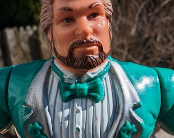 May include: A green and silver action figure of a man wearing a tuxedo with a green bow tie and dollar sign embellishments on the jacket.