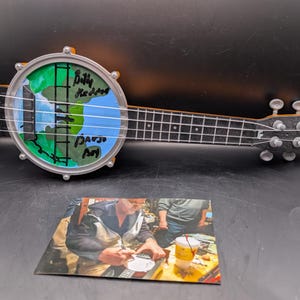 May include: A banjo ukulele with a painted world design on the drumhead. The artwork includes green landmasses and blue oceans, with the text "Billy Redden" and "David M" written vertically. The instrument has a black neck and silver hardware.