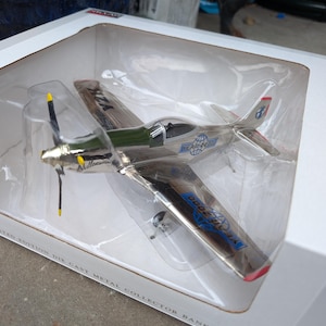 Puede incluir: Un avión modelo de colección de metal fundido a presión en una caja de plástico transparente. El avión es plateado con detalles en verde y azul, una cabina negra y una hélice con punta amarilla. La caja dice "LIMITED EDITION DIE CAST METAL COLLECTOR BANK."