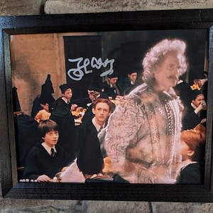 以下が含まれることがあります： ハリーポッター映画シリーズのシーンが写った額縁付きの写真。白いローブを着たキャラクターと、黒いローブを着た生徒たちのグループが写っている。写真は「J.K. ローリング」のサイン入りです。