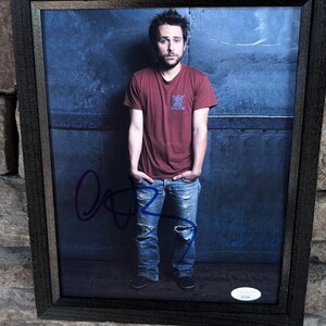 Può includere: Una fotografia in bianco e nero incorniciata di un uomo che indossa una maglietta rossa e jeans blu. L'uomo è in piedi davanti a un muro blu e ha una firma in inchiostro blu sulla foto. La firma recita "Rob McElhenney".