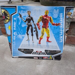 May include: A Marvel Legends action figure set featuring Iron Man and Maria Hill. Iron Man is wearing a red and gold suit of armor. Maria Hill is wearing a black suit with a gun and a backpack. The box features a comic book style background with the Marvel Legends logo.