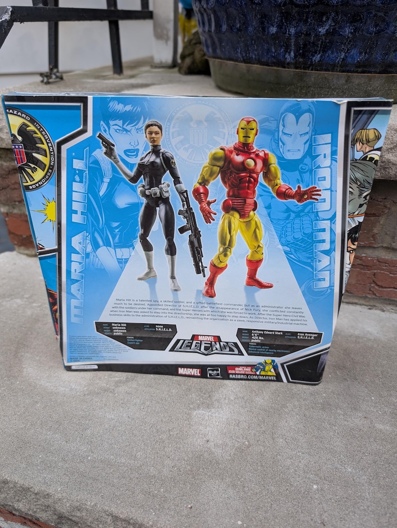 May include: A Marvel Legends action figure set featuring Iron Man and Maria Hill. Iron Man is wearing a red and gold suit of armor. Maria Hill is wearing a black suit with a gun and a backpack. The box features a comic book style background with the Marvel Legends logo.