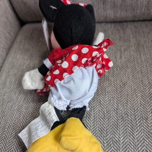 K&ouml;nnte beinhalten: Ein Minnie Mouse Pl&uuml;schtier, mit einem roten Kleid mit wei&szlig;en Punkten, einer passenden Schleife und gelben Schuhen. Die schwarzen Ohren und das Gesicht sind sichtbar. Das Spielzeug liegt auf einer grauen Oberfl&auml;che.